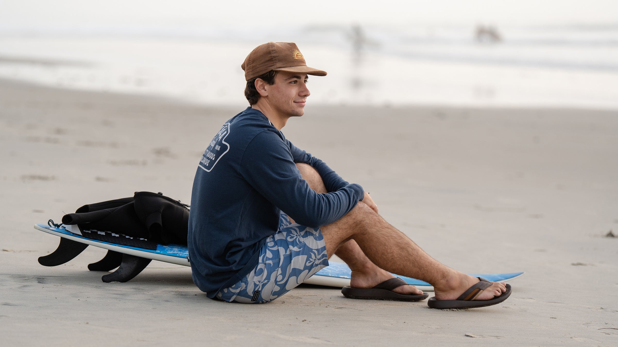 A man sitting on the sand next to a surfboard, wearing Cobian Kandui Chocolate Sandals