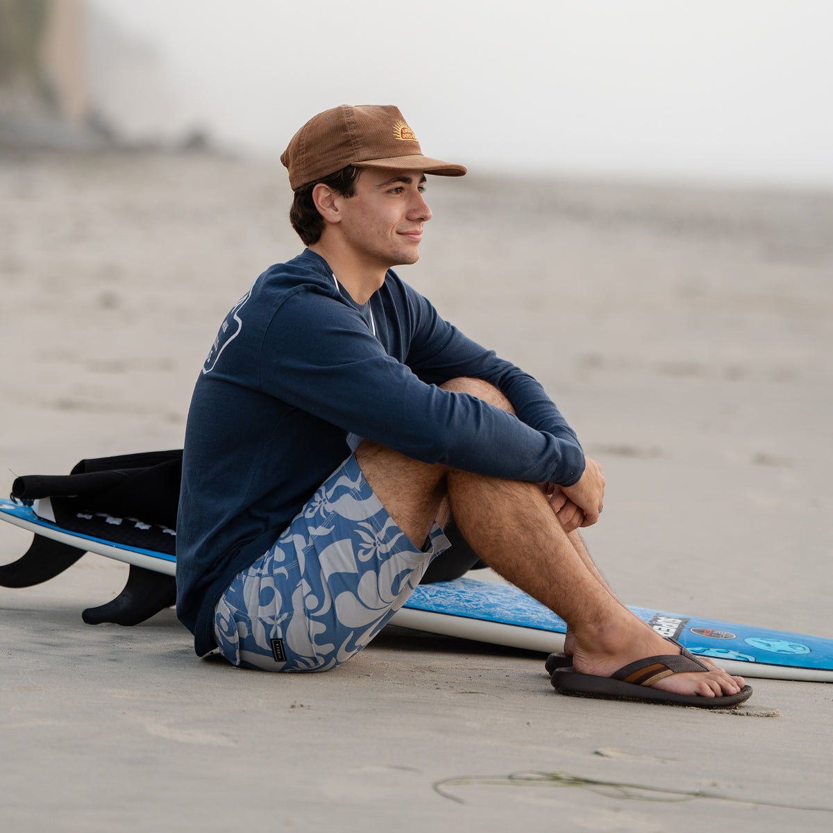 A man sitting on the sand next to a surfboard, wearing Cobian Kandui Chocolate Sandals