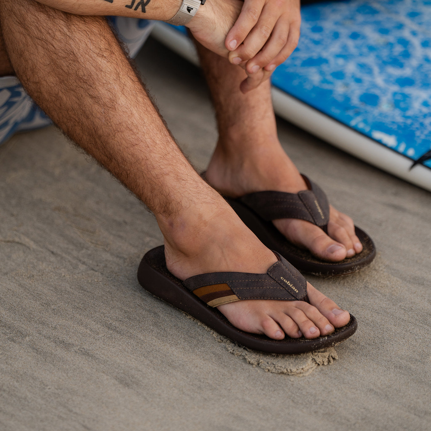 A close-up of a man wearing the Cobian Kandui Chocolate Sandals on a sandy beach #color_chocolate